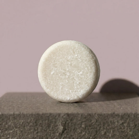 Round, light-colored shampoo bar on a textured surface with a soft pink background