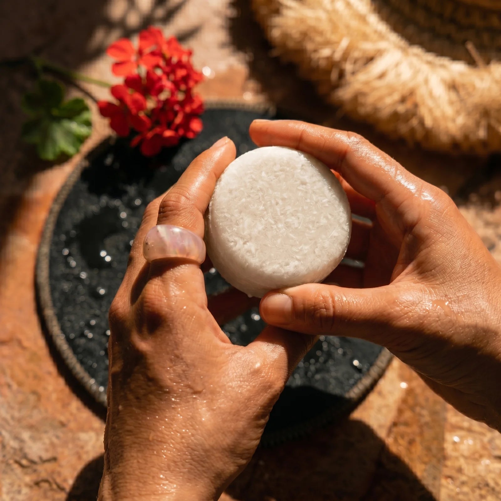 Person holding a white shampoo bar with a red flower and a straw hat in the background